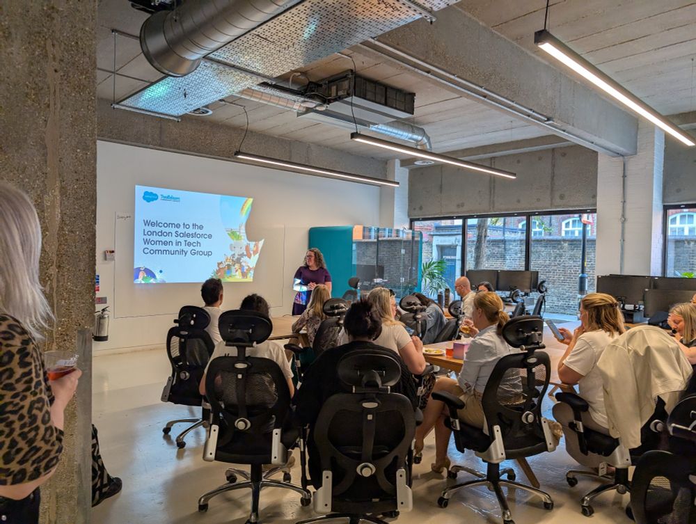 A woman stands in front of a presentation screen displaying the title "Welcome to the London Salesforce Women in Tech Community Group" in a modern office setting. Attendees, mostly women, are seated and listening attentively, with some enjoying refreshments.