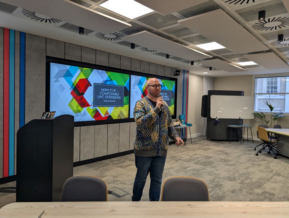 A speaker stands in front of a presentation slide titled "Mixin It Up - Composable LWC Extensions" by Diego Cominazzini. He is holding a microphone in a modern office-style room with colourful wall panels, a large screen, and various seating arrangements.
