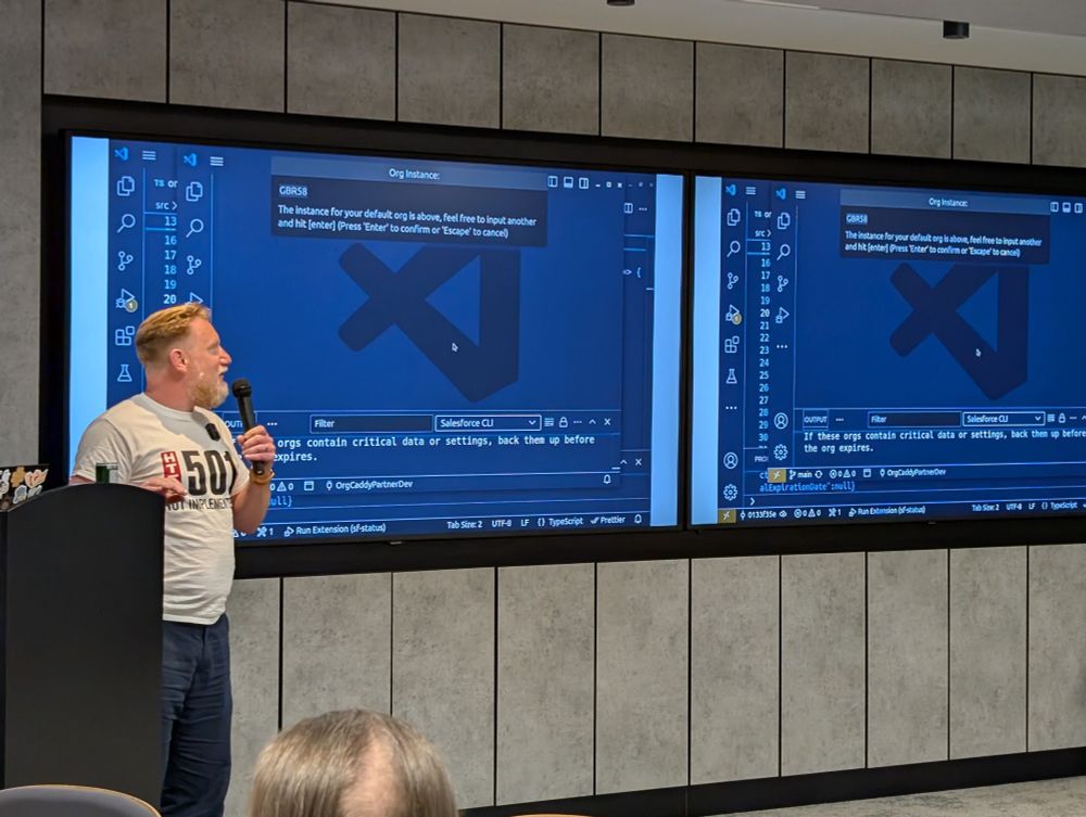 A man is speaking into a microphone while standing in front of a podium during a presentation. Behind him are two large screens displaying Visual Studio Code with a Salesforce CLI terminal open. He is wearing a grey t-shirt with a bold red and black logo and text that says "HTTP 501 NOT IMPLEMENTED"
