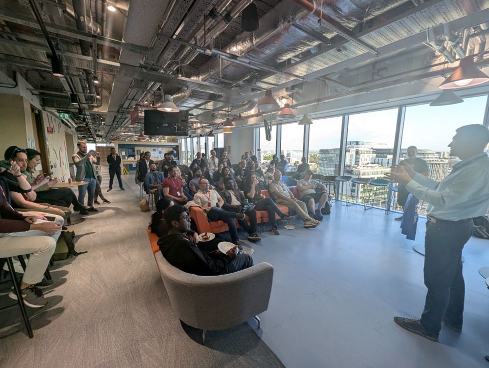 A larger audience is seated and standing in a bright, open-plan office, listening attentively to the speaker from the first image. The room is filled with natural light from the expansive windows overlooking central London, with a mix of attendees enjoying food and taking photos.