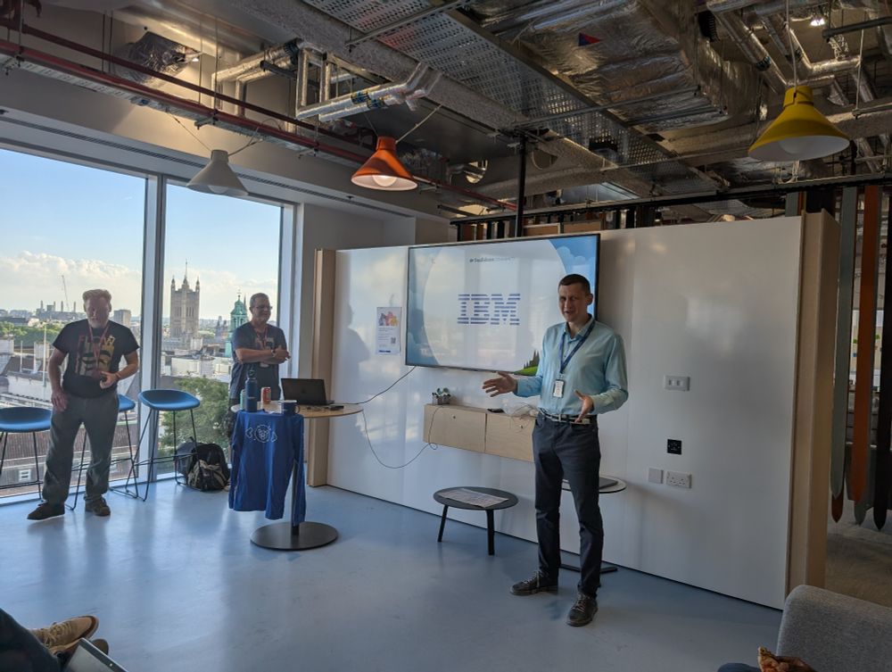 A speaker stands in front of a screen displaying the IBM logo, addressing a small group during a tech meetup. Two other individuals stand nearby, one behind a table with merchandise and drinks. The event takes place in a modern office with large windows offering a clear view of Westminster, including the Victoria Tower.
