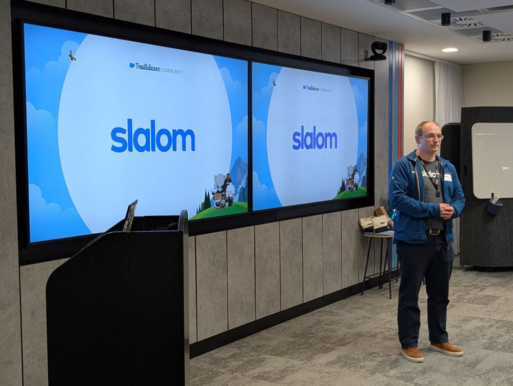 A man wearing a Slalom-branded jacket and t-shirt is standing and speaking in front of two large screens displaying the Slalom logo and the Salesforce Trailblazer Community branding. The event appears to be taking place in a modern office or conference room. A lectern and some branded merchandise are also visible.