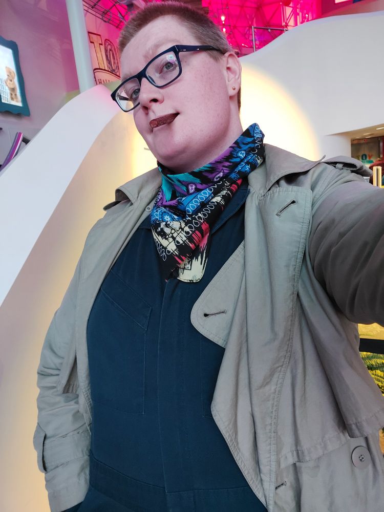 Picture of the OP, Talia, posing at a museum, wearing a jumpsuit, trenchcoat, and rainbow bandana, holding a short slimjim meat snack in her mouth like a cigar 