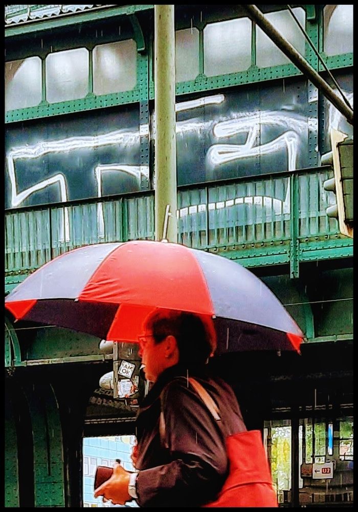 Farbaufnahme an einem Regentag. Eine Frau, von der nur die obere Körperhälfte sichtbar ist und die einen großen Regenschirm trägt, läuft eilig an einer Hochbahnstrecke entlang, während sie einen Schulterbeutel trägt.