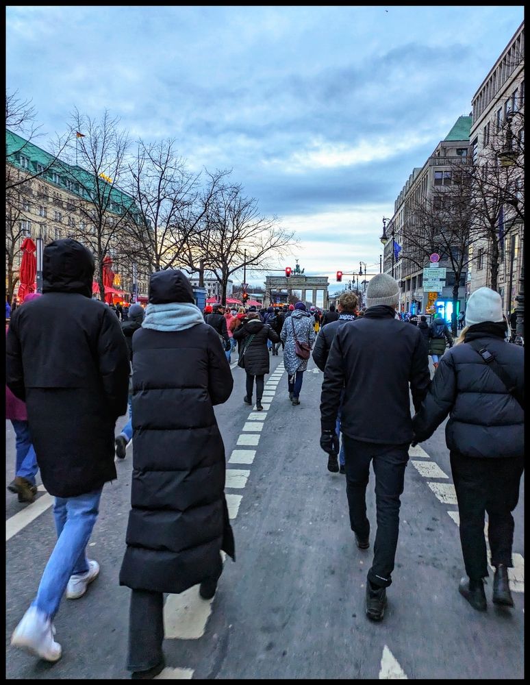Farbaufnahme in eine beginnende Demo. Auf einem Boulevard strömen Menschen zum Brandenburger Tor. Winterende.