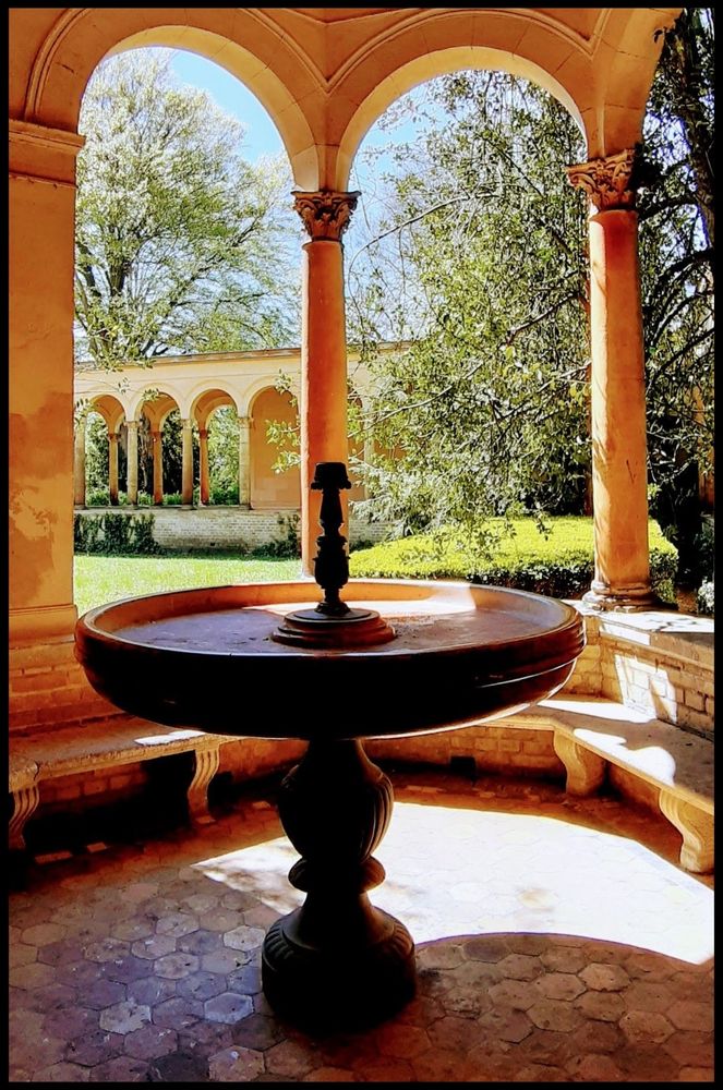 Farbaufnahme eines angelegten Hofes einer Kirche. Blick durch Kolonnaden auf einen grünen Innenhof. Im Vordergrund eine Wasserschale als Standbrunnen. Terracotta herrscht vor.