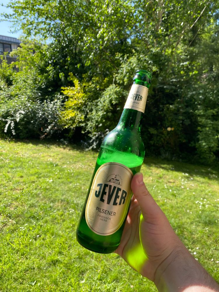 My hand holding a bottle of Jever in a green, sunny garden 