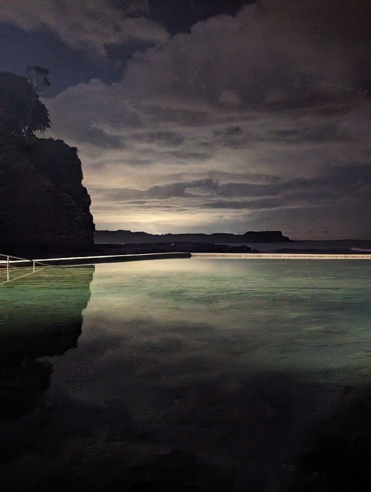 A long exposure night time photo of a coastal rock pool, with clouds in the sky & a mirrored reflection visible on the surface of the water