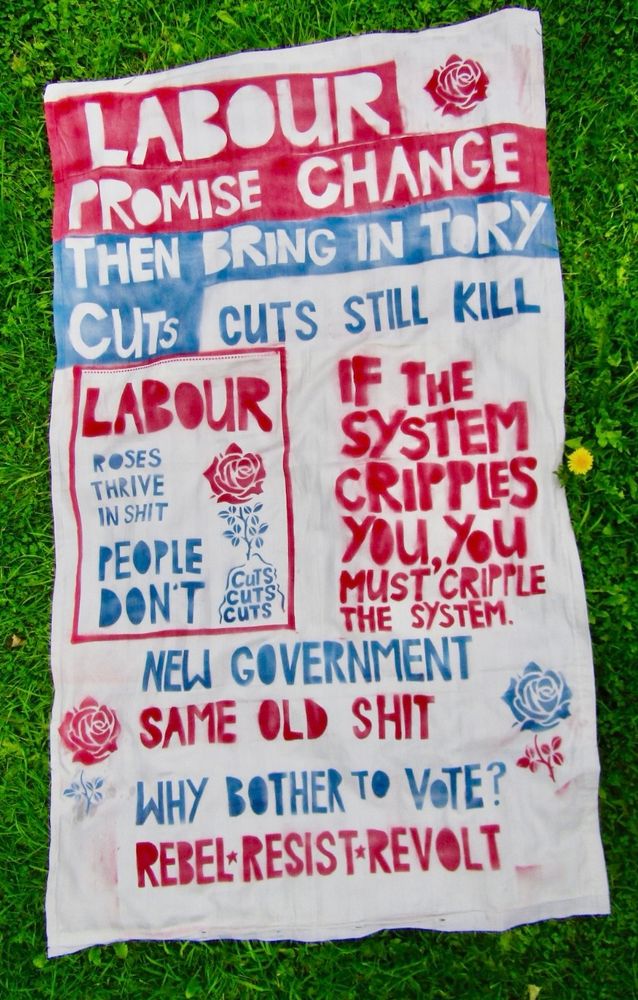 
Labour 
promise change
then bring in Tory cuts
cuts still kill
Labour
Roses thrive in shit
People don’t. Cuts cuts cuts.
If the system cripples you
you must cripple the system.
New government
same old shit
Why bother to vote?
Rebel Resist Revolt

The shroud is white fabric with red and blue text, with red and blue roses.