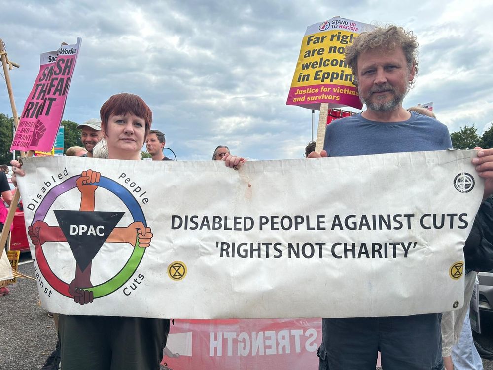 Woman with short red hair wearing black stands next to a man wearing dark blue with curly dark hair and a grey beard holding a white banner with DPAC Colour Logo with black text Disabled People Against Cuts Rights Not Charity 