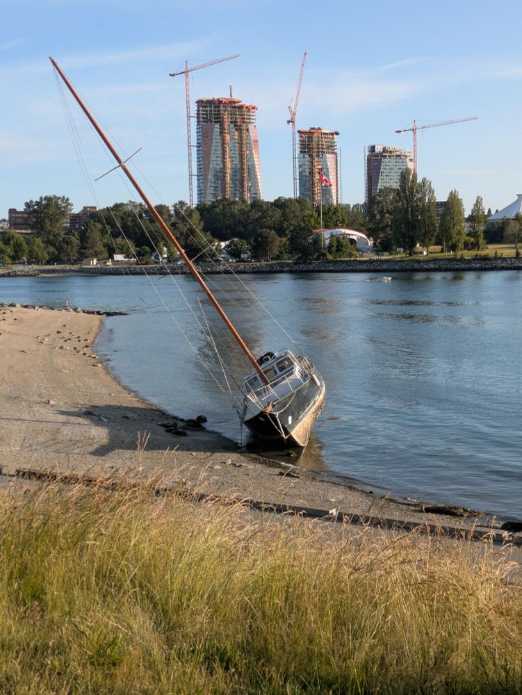 A boat washed up on the shore with towers under construction across the water in the distance 