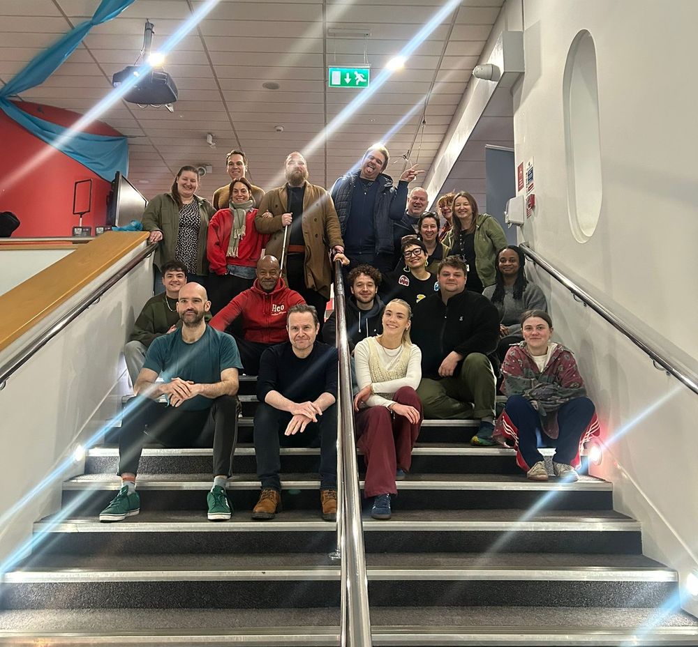 A group photo of around 20 people sitting on a staircase in a brightly lit indoor space. The group appears cheerful, with a mix of casual and winter attire, and blue fabric decorations hanging from the ceiling.