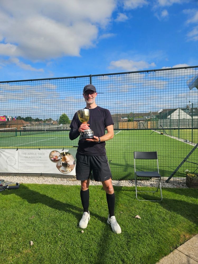 A middle aged man in black hat,  t shirt, shorts and socks holding a trophy in front of a tennis court