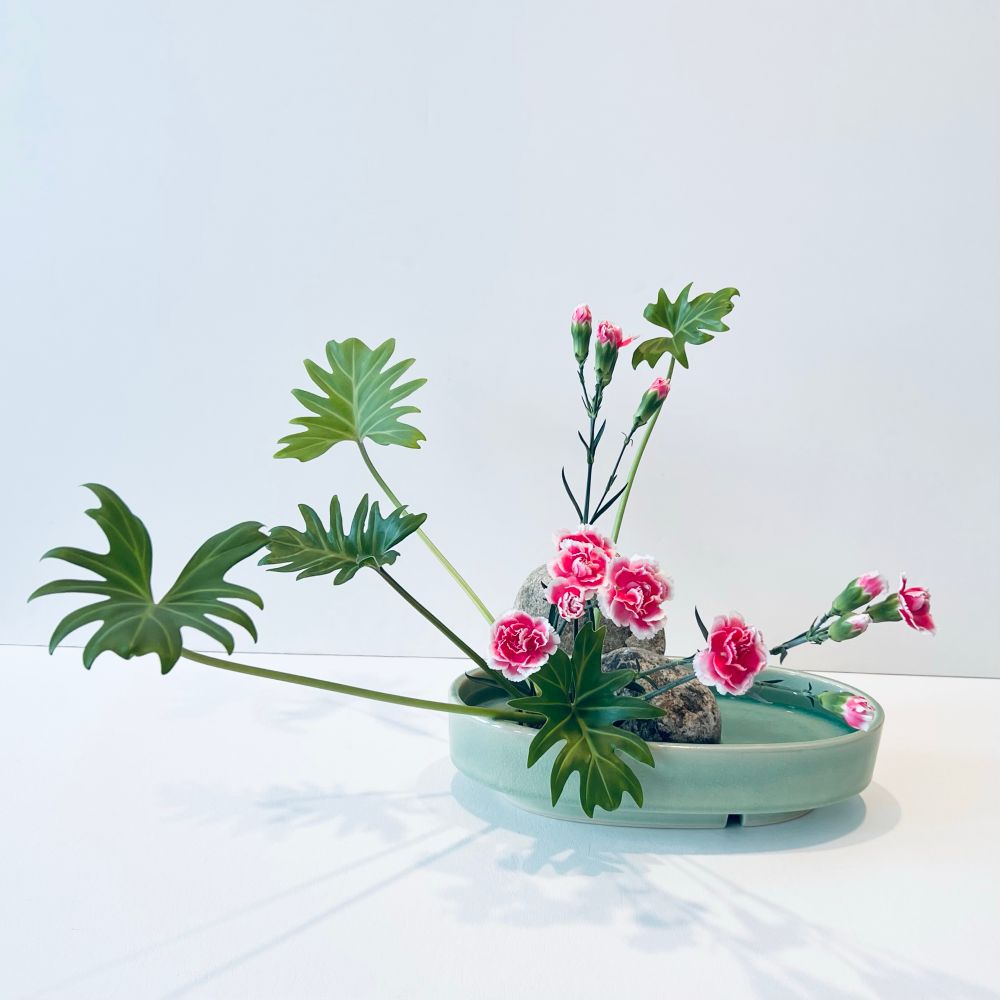 Ikebana arrangement in an celadon oval container. Flowers include Xanadu and pink and white carnations. Rocks in background