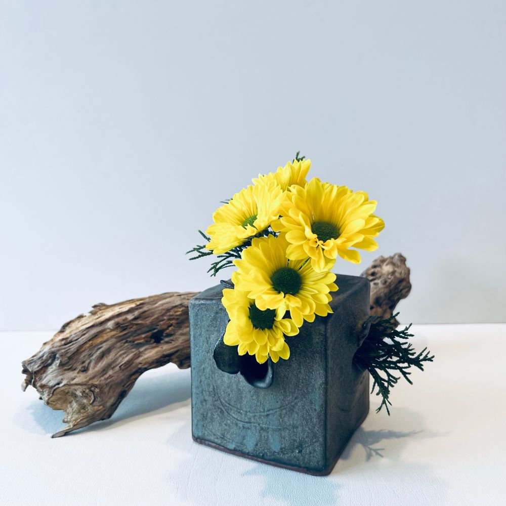 Bright yellow mums and pine in a green square vase. Driftwood in the background.