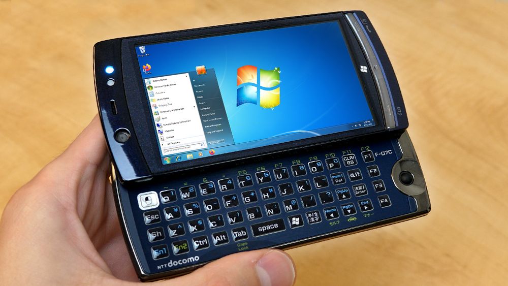 a YouTube thumbnail featuring a phone running Windows 7 being held above a wooden table. On screen is the default Windows 7 desktop wallpaper, with the start menu open.