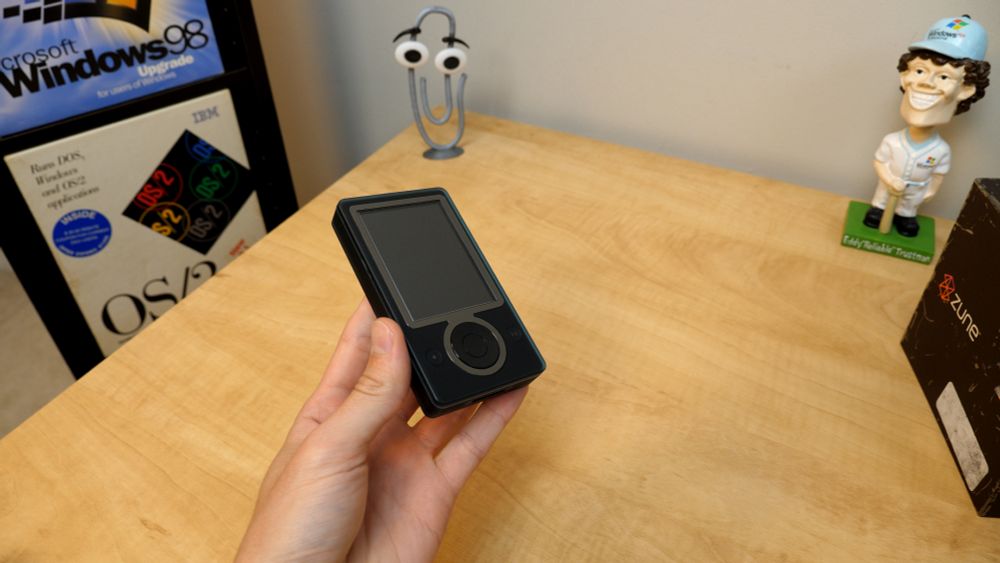 a Microsoft Zune being held above a wooden table. Visible to the right is the Zune box, along with the Eddy "Reliable" Trustman bobble-head. There is a 3D printed Clippy mascot is near the top of the image. To the left is a shelf that has boxed copies of Windows 98 and OS/2.