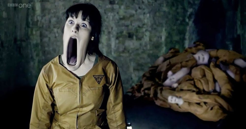 Scary scene from the Doctor Who episode The Almost People. A plastic ‘flesh’ doppelgänger is staring at the camera with her lower jaw elastically drooping downwards. Behind them are discarded ‘bodies’ of flesh copies. 