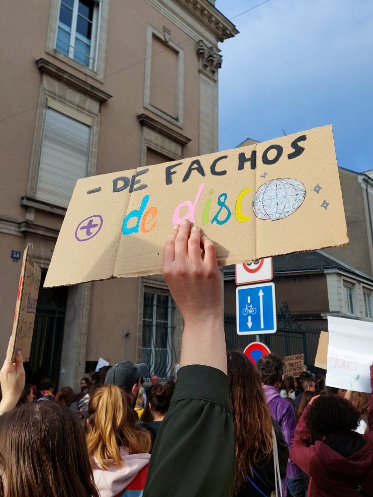 Dans la foule, une personne tient d'une main, une pancarte en hauteur avec le slogan : « Moins de fachos plus de disco ».