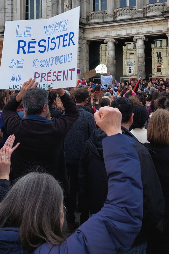 Dans la foule, une femme dresse un point serré devant une pancarte citant Lucie Aubrac : « Le verbe resister se conjugue au présent ».