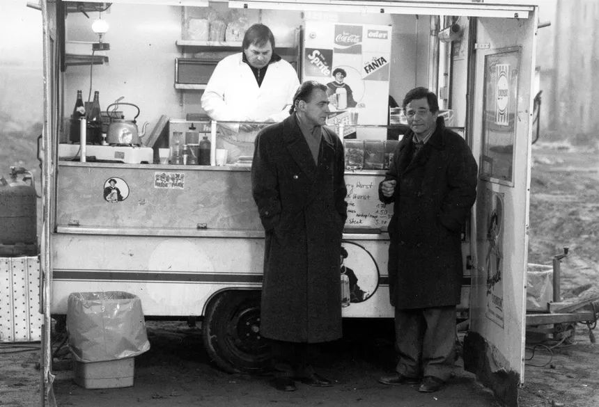 Les acteurs Bruno Ganz et Peter Falk en pleine discussion adossés à un stand de restaurant mobile sur un terrain vague.