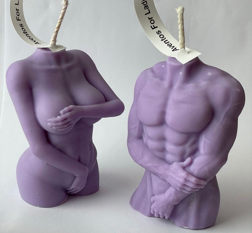 Two naked torso figure candles of a lilac colour. The one on the left is a naked woman covering her vagina and nipples with her hands and the one on the right is a muscly naked man holding one arm 
