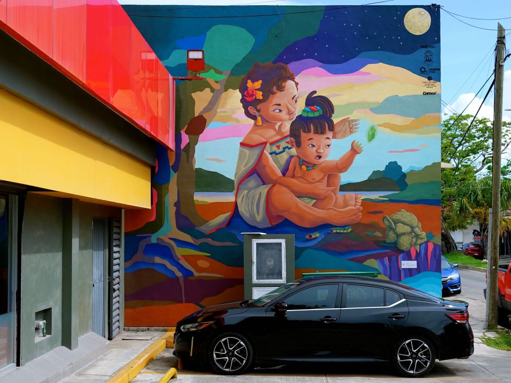 Ein großflächiges buntes Wandgemälde (Mural) an einer Gebäudewand in Chetumal zeigt eine Maya-Frau mit Blume im Haar, die ein kleines Kind auf dem Schoß hält. Die Szene ist in leuchtenden Farben gemalt - mit einem sternenklaren Nachthimmel, Vollmond, tropischer Landschaft und einem Ceiba-Baum im Hintergrund. Die Frau trägt traditionelle Maya-Kleidung mit bunten geometrischen Mustern. Verschiedene Maya-Symbole wie eine Schlange und ein Jaguar sind in das farbenfrohe Kunstwerk integriert. Ein schwarzer Pkw steht vor dem Mural des Künstlers Dherzu Uzala, das Teil des Projekts "Rutas Mágicas de Color" ist.