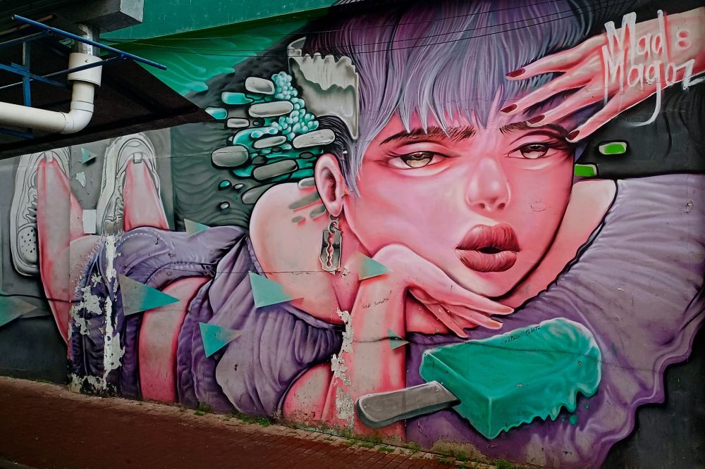 Lebendiges Wandbild in Santa Ana, Panama-Stadt, das ein stilisiertes Porträt einer Person mit violettem Haar und rosa-getönter Haut zeigt. Die Figur trägt eine violette Jacke und hat die Hand nachdenklich nah am Gesicht positioniert. Das Kunstwerk enthält surreale Elemente wie schwebende pillenförmige Objekte in Türkis und Grau sowie fließende organische Formen. Das Werk ist in der oberen rechten Ecke mit "Mad Magos" signiert. Das Wandbild ist auf eine Betonwand mit sichtbaren Versorgungsleitungen gemalt, und am unteren Rand der Komposition befindet sich eine Betonkante.