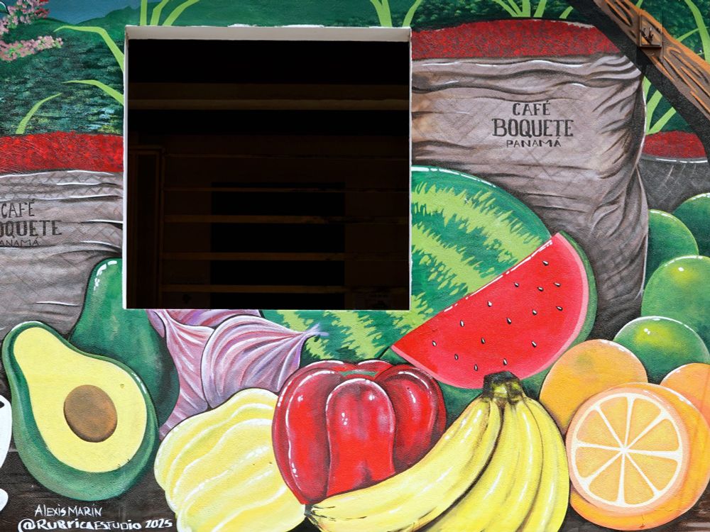Nahaufnahme des unteren Bereichs des Murals mit einer lebendigen Darstellung tropischer Früchte und Produkte: eine aufgeschnittene Avocado, rote Paprikaschoten, Bananen, eine Wassermelonenscheibe, Orangen und Kaffeesäcke mit der Aufschrift "Café Boquete Panama". Die Signatur des Künstlers "Alexis Marin @RubricaEstudio 2025" ist in der unteren linken Ecke sichtbar.