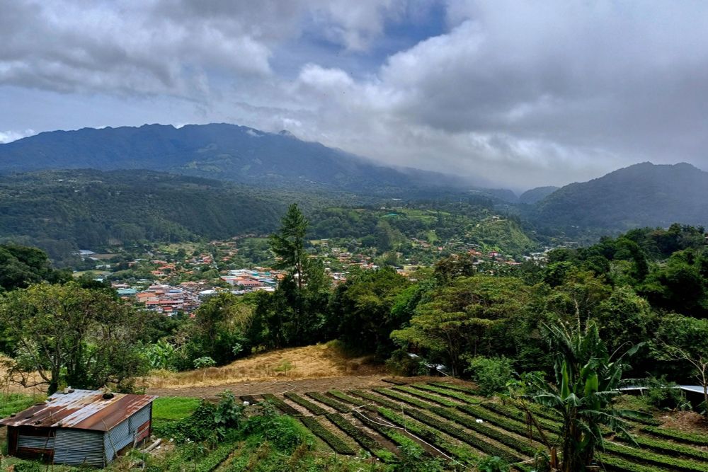 Panoramablick von der Straße zwischen Jaramillo und Boquete auf das Tal in der Provinz Chiriquí: Im Vordergrund sind terrassierte Gemüsefelder und ein Wellblechhaus zu sehen, dahinter erstreckt sich die Ortschaft Boquete mit ihren roten Ziegeldächern im grünen Talkessel. Die Landschaft ist geprägt von dicht bewaldeten Berghängen und dramatischen Wolkenformationen über den hohen Gipfeln der Cordillera de Talamanca. Der bewölkte Himmel und das saftige Grün der Vegetation zeigen das typische Klima der panamaischen Bergregion.