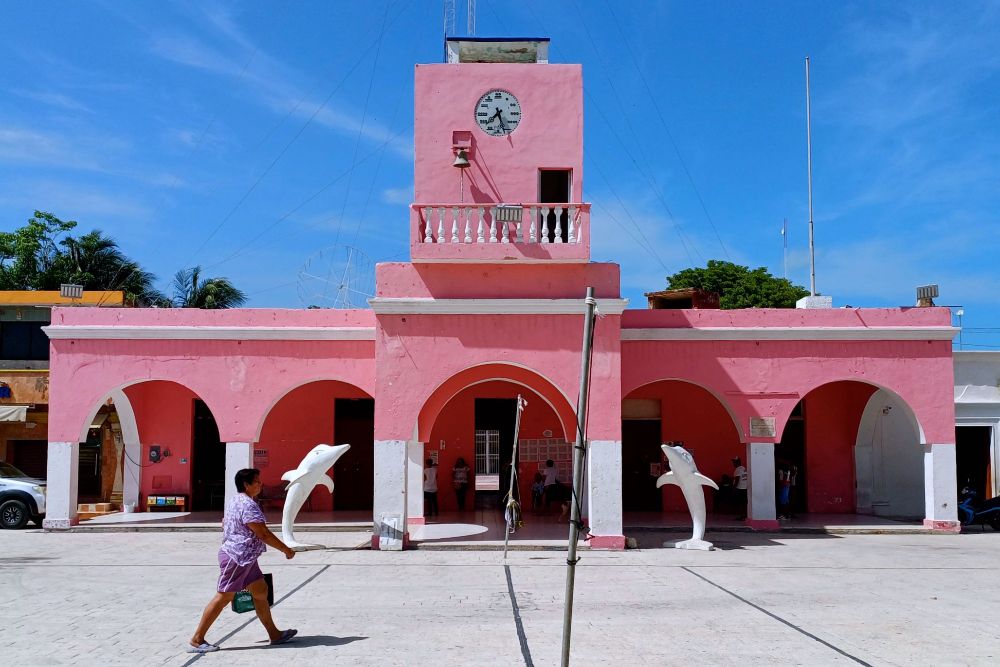Der Palacio Municipal de Celestún in leuchtend rosa-roter Farbe mit weißen Akzenten. Das zweistöckige Kolonialgebäude hat fünf Rundbögen im Erdgeschoss und einen Uhrenturm mit weißem Balkon. Eine Uhr ist an der Turmfront sichtbar. Vor dem Gebäude überquert eine Frau in violettem Kleid den gepflasterten Platz. Im Vordergrund stehen zwei weiße Delfinskulpturen als Dekoration. Der Himmel ist strahlend blau mit wenigen weißen Wolken.