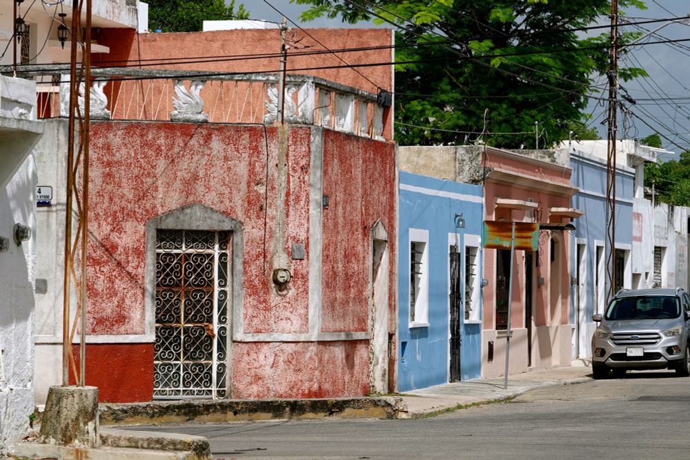 Straßenszene in Mérida mit einer Reihe bunter Kolonialhäuser - rot mit ornamentalem Eisengitter, hellblau und rosa - entlang einer gepflasterten Straße mit einem geparkten silberfarbenen Fahrzeug.