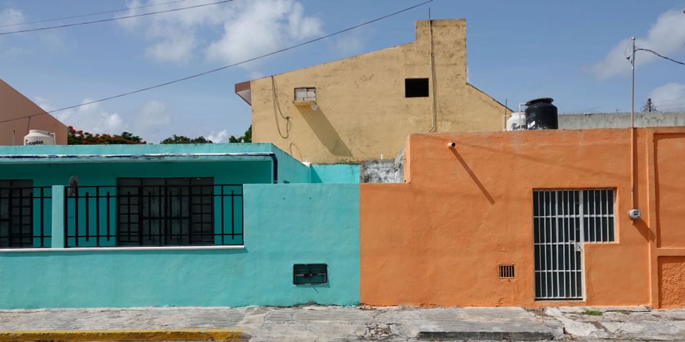 Bunte Wohnhäuser in Mérida mit türkisfarbener und orangefarbener Fassade nebeneinander, beide einstöckig mit vergitterten Fenstern, dahinter ein beiges mehrstöckiges Gebäude unter blauem Himmel mit weißen Wolken.