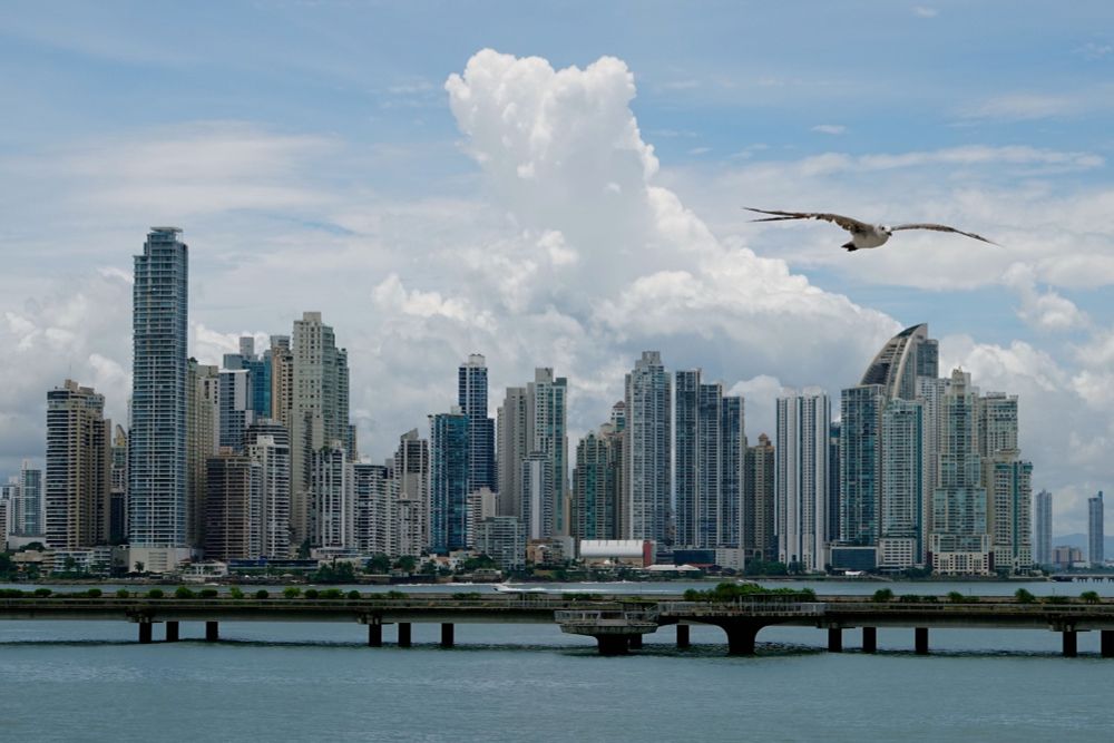 Panoramablick auf die moderne Skyline von Panama-Stadt mit zahlreichen Hochhäusern und Wolkenkratzern in verschiedenen Höhen. Die Gebäude zeigen eine Mischung aus Glas- und Betonfassaden in Blau-, Grau- und Beigetönen. Im Vordergrund erstreckt sich die Bucht mit ruhigem Wasser, über die eine niedrige Brücke mit mehreren Pfeilern führt, die zur Cinta Costera 3-Infrastruktur gehört. Eine Möwe fliegt mit ausgebreiteten Flügeln durch das Bild. Der Himmel zeigt dramatische weiße Kumuluswolken vor einem blauen Hintergrund. Am gegenüberliegenden Ufer ist grüne Vegetation zu sehen.