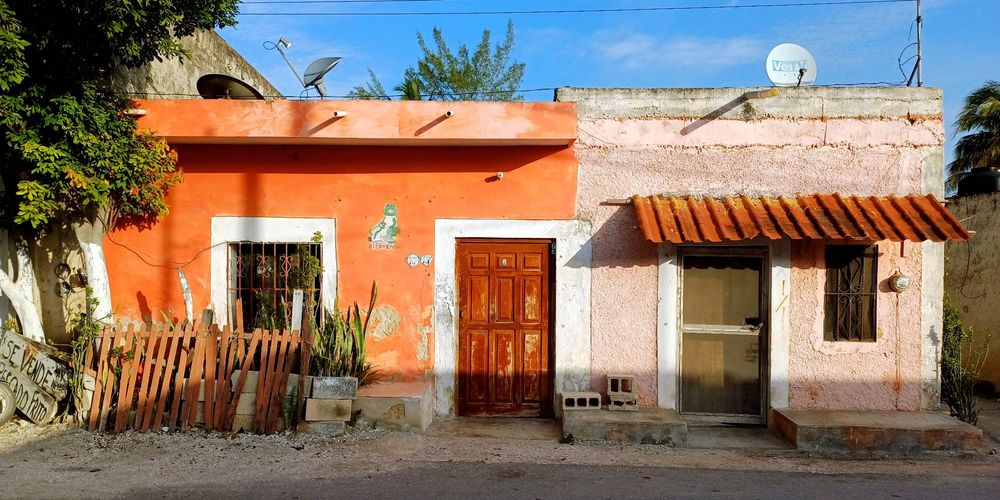 Charakteristische mexikanische Wohnhäuser in Celestún in lebendigen Farben. Das linke Haus ist leuchtend orange gestrichen mit weißen Fensterrahmen und einer braunen Holztür. Rechts daneben steht ein rosa-beiges Haus mit roten Ziegeln als Vordach. Vor den Häusern wachsen Agaven und andere tropische Pflanzen hinter einem hölzernen Lattenzaun. Satellitenschüsseln und Stromleitungen sind auf den Dächern sichtbar, unter einem strahlend blauen Himmel.