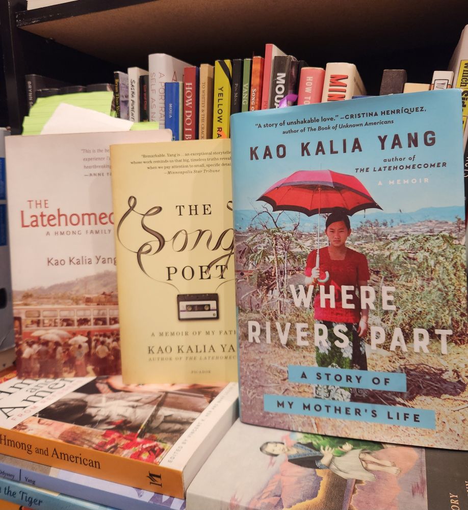 Three memoirs by Kao Kalia Yang standing against a shelf of books. From left to right, the books are "The Latehomecomer," "The Song Poet," and "Where Rivers Part."