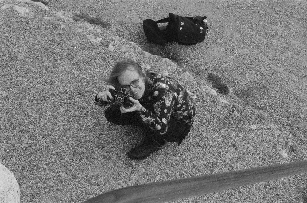 A girl crouches on the ground taking a photo up towards the elevated point of view of the photo. She is using a folding camera from the 1930s. The photo is in black and white film. 