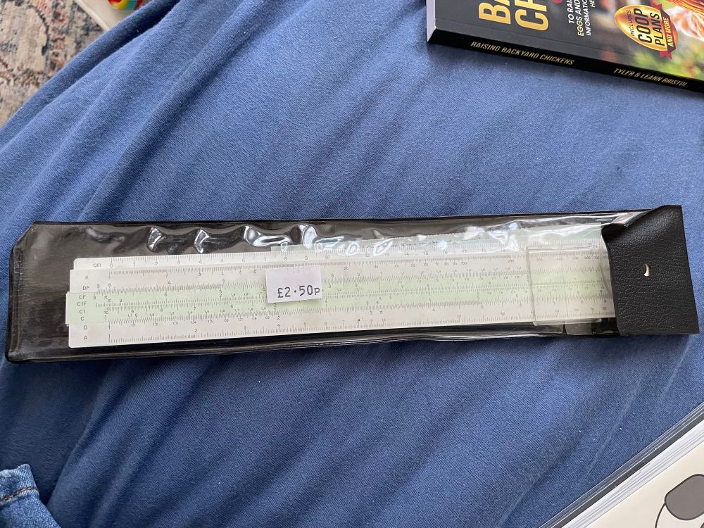 A white and green slide rule (a ruler with sliding bar used to calculate logarithmic and quadratic equations) in original packaging on a blue background. An attached price label says £2.50p. 