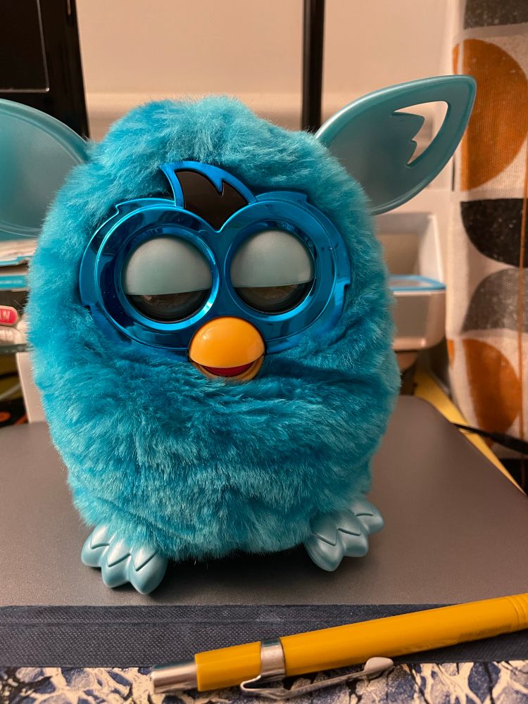 A bright blue furry toy with big round eyes like an owl, a small yellow beak and big plastic ears like a gremlin or elf, sits on a desk next to a notepad and pen. The eyes are half closed. 