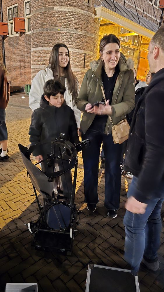 Two young women are talking to a man (with his back to the camera) while a young boy is looking at the man's telescope. Behind them is a turret from the Waag on the nIeuwmarkt. 
