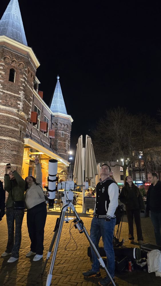 A young man is standing next to a telescope, the telescope is pointed up and the man is looking up as well. Behind him there are other people looking up and pointing at the sky. Behind them all is the Waag of the Nieuwmarkt in Amsterdam. 