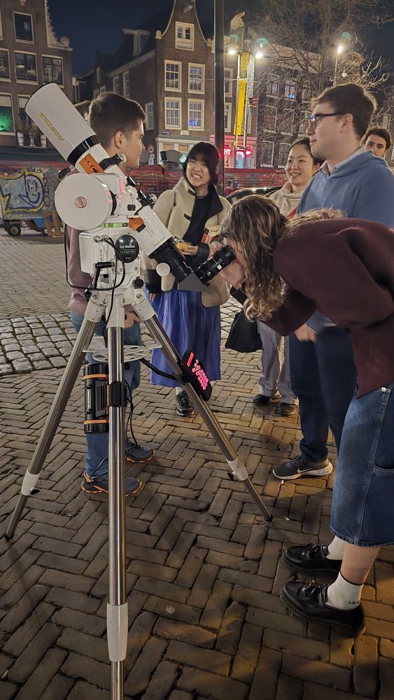 A woman is looking through a telescope. Behind her, a few people are smiling and chatting. In the background you see typical Amsterdam architecture. 