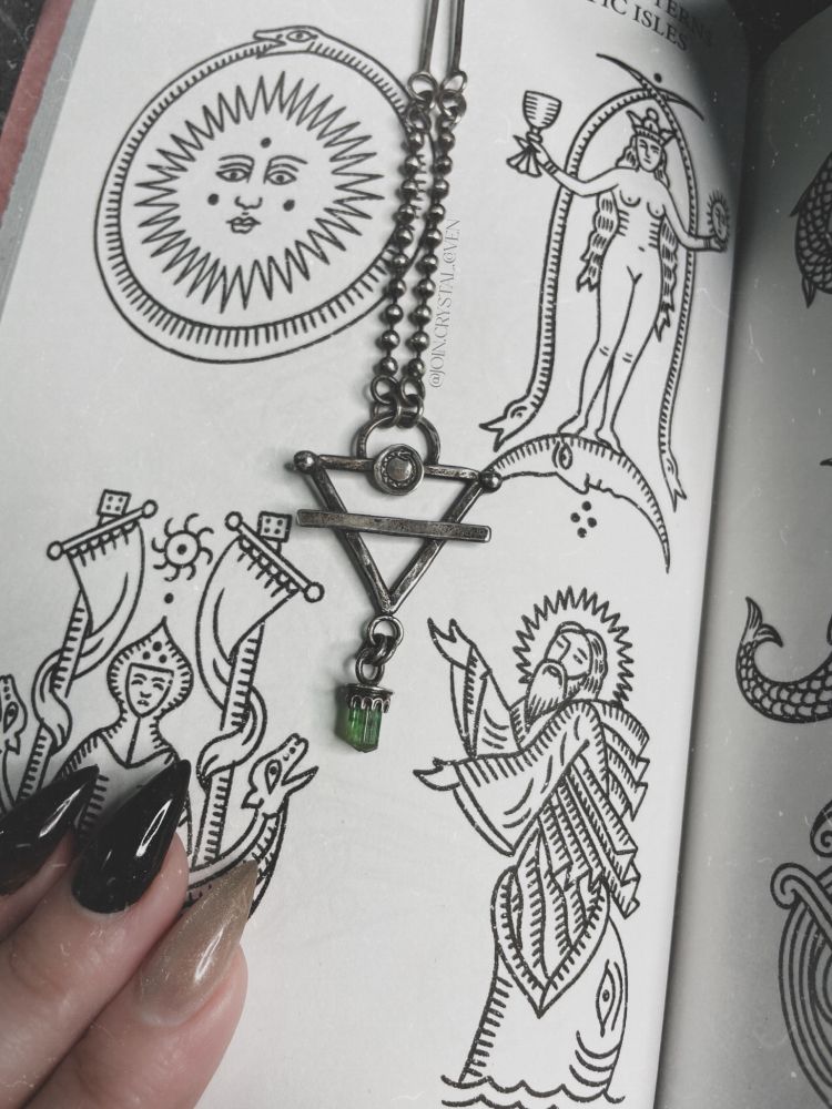 A handmade sterling silver and green tourmaline talisman necklace lays on an open book depicting Celtic patterns and designs. The necklace is forged into the shape of the earth alchemy symbol (upside down triangle with a line through it), with a small ouroboros sitting at the top and a green tourmaline terminated crystal hanging down from the bottom.