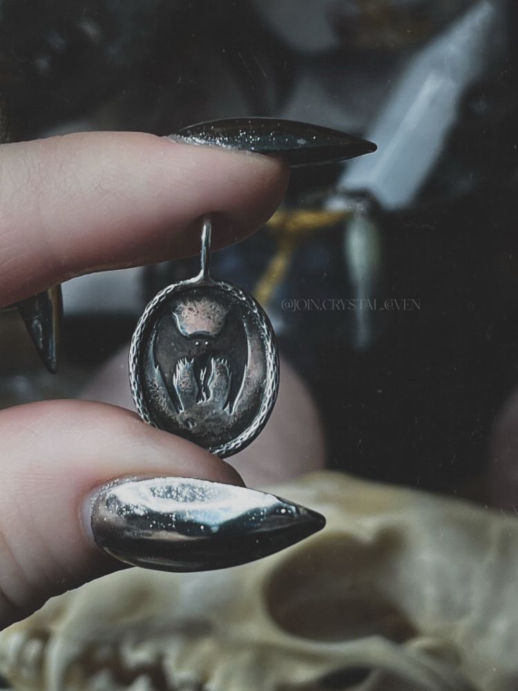 Witchy hands with long silver chrome nails holds an oval Mothman pendant. The pendant is made of sterling silver and the silhouette of Mothman is blackened for contrast.