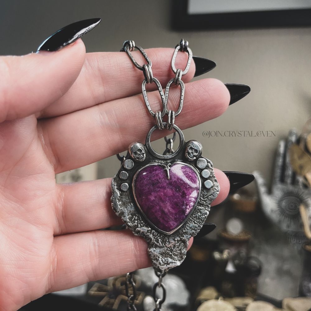 A hand holds a sterling silver necklace with a large ruby heart surrounded by two skulls and 6 circles. The bottom of the necklace looks intentionally melted to give texture.