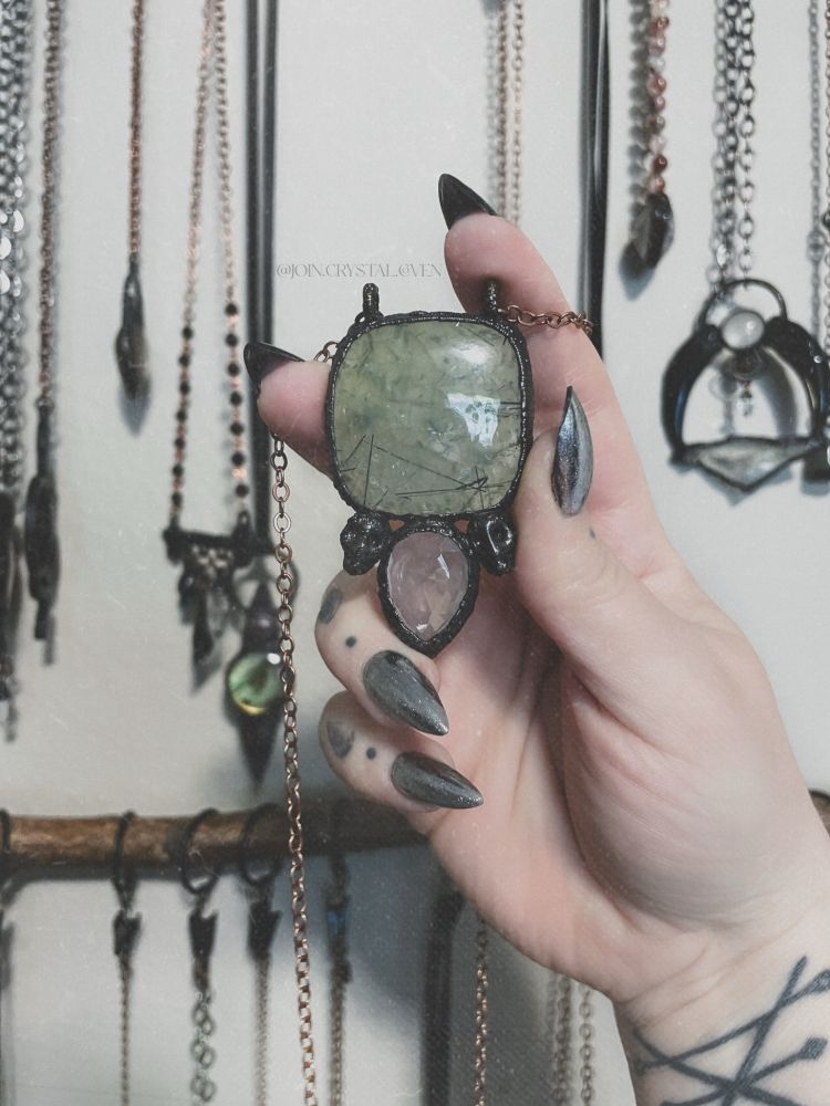 A hand holds a large necklace featuring a square with rounded edges large green Prehnite atop a faceted pink Rose Quartz upside down teardrop with a small skull on other side. The metal is blackened copper.