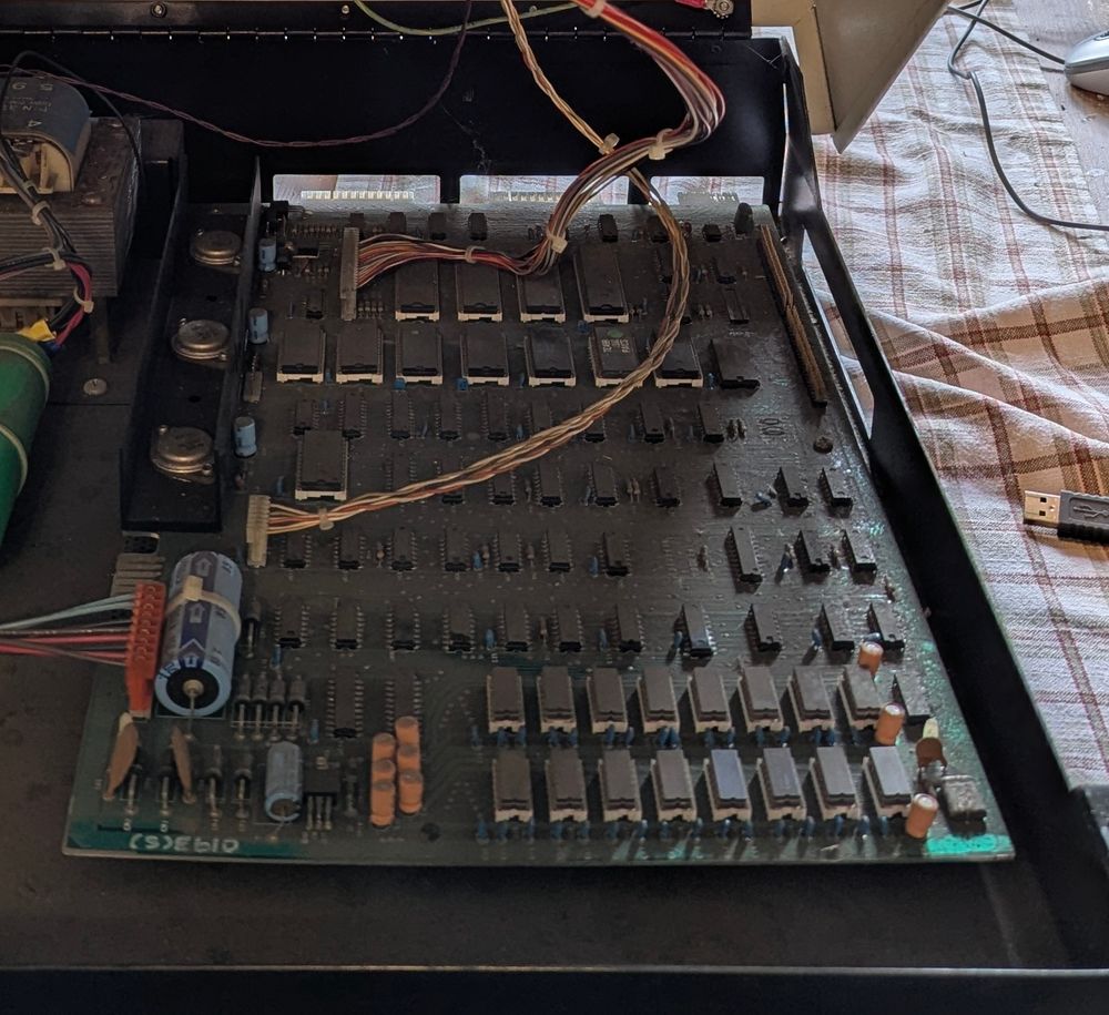 A picture of the Commodore PET motherboard. It's very dirty. There are sixteen RAM chips in white sockets. Seven ROM chips are also socketed towards the back. A large blue capacitor is towards the front left of the board and there are three round voltage regulators behind the capacitor. 