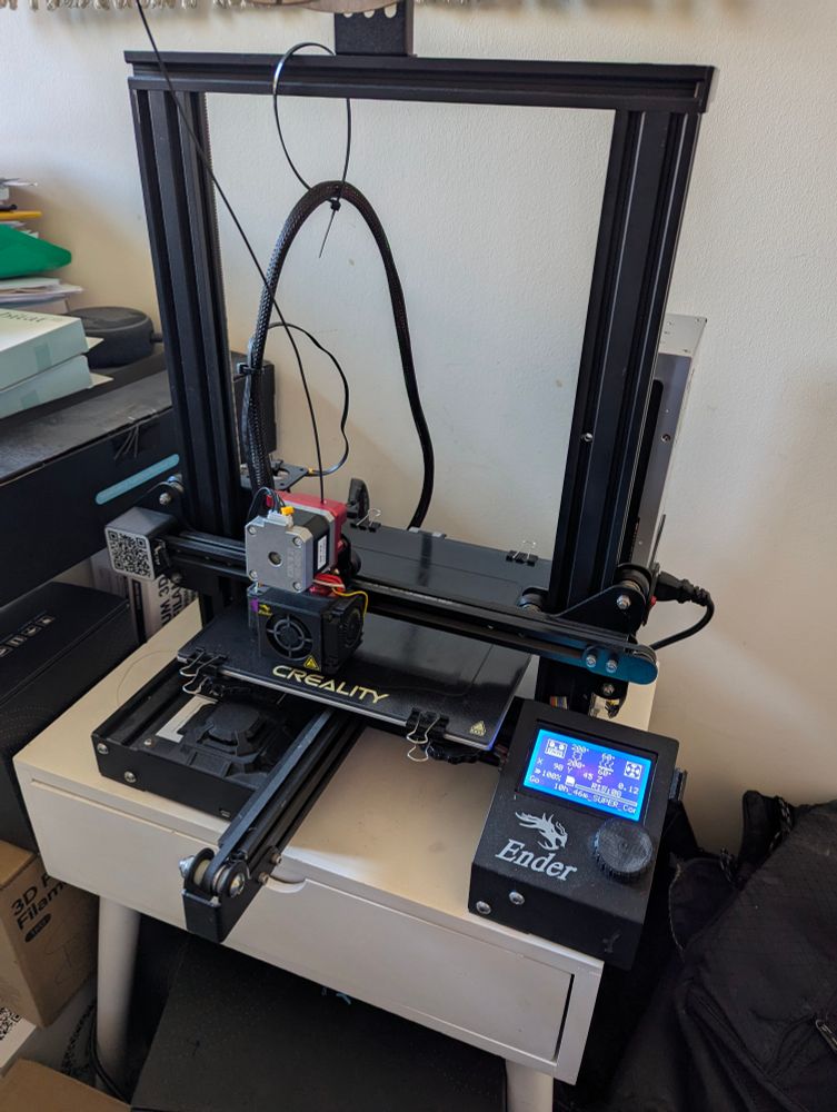 A picture of an Ender 3 3D printer with direct drive and red filament feed mechanism. The printer is printing black filament onto a glass bed. The printer is running firmware from Tiny Machines.