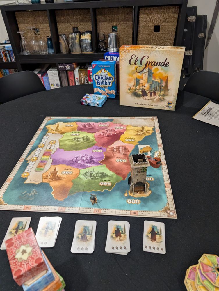 Board game El Grande set up on a black table with shelves of games and cups in background.