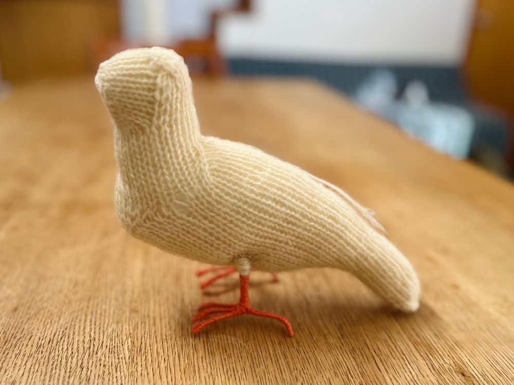 白色の毛糸で編まれたシンプルな鳥。オレンジがかったピンク色の糸が巻かれた細い足がついている。
